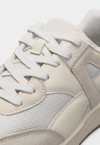 Μπεζ και λευκό αθλητικό παπούτσι με πάνελ από δίχτυ και σουέτ, με λευκά κορδόνια και επενδυμένο γιακά για άνεση και υποστήριξη.