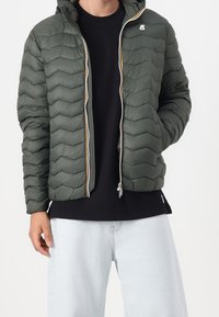 Homme portant une veste matelassée olive verde dézippée avec capuche, un t-shirt noir et un jean bleu clair, se tenant devant un fond uni.
