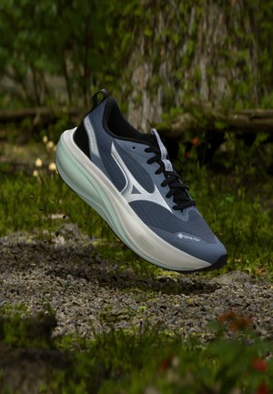 Blå og sort løbesko med hvid sål og Gore-Tex mærkat, ophængt over en grussti i en skov.