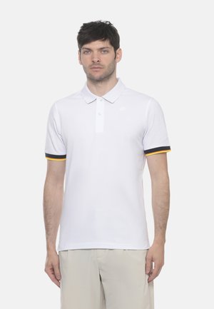 Uomo in piedi rivolto in avanti, indossa una polo bianca con rifiniture nere e gialle sulle maniche e pantaloni beige su sfondo chiaro.