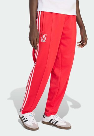 Person trägt rote Trainingshose des Liverpool Football Clubs mit weißen Streifen an den Seiten und weiße Adidas-Sneaker mit schwarzen Streifen.