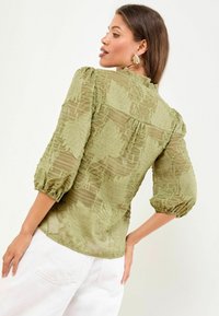 Blusa verde oliva con motivi di foglie testurizzati, maniche a palloncino e scollatura rotonda. Tessuto trasparente con dettagli arricciati sulle spalle.