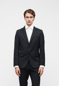 Blazer azul-marinho com dois botões, lapelas notched e um único bolso no peito, combinado com uma camisa branca de botões e calças escuras.