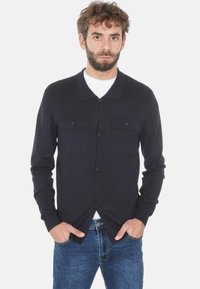 Uomo con barba che indossa un cardigan blu scuro con bottoni sopra una camicia bianca e jeans blu, con le mani in tasca, rivolto in avanti su uno sfondo bianco.