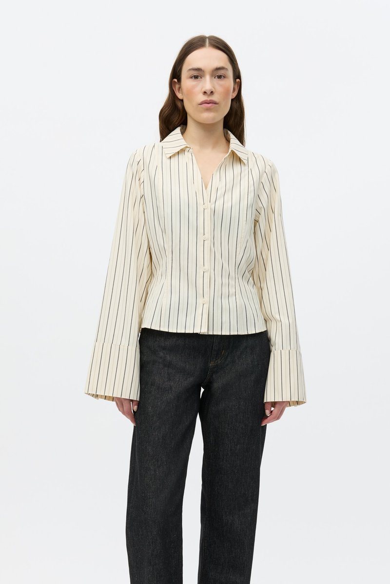 Ριγέ πουκάμισο με κουμπιά σε ανοιχτό κρεμ και μαύρο χρώμα, με V-λαιμόκοψη, μακριά φαρδιά μανίκια και εφαρμοστή μέση. Συνδυάζεται με σκούρο τζιν.