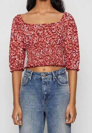 Cropped top floreale rosso con maniche a sbuffo e scollatura quadrata; texture arricciata in vita. Abbinato a jeans a vita alta blu sbiaditi.