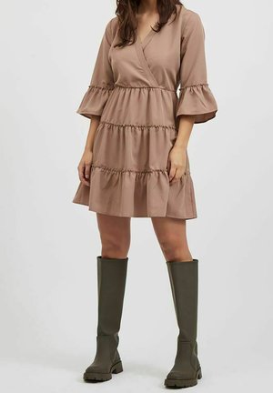Lichtbruine wikkeljurk met gelaagde ruches, lange klokkende mouwen en een getextureerde afwerking; gecombineerd met kniehoge olijfgroene laarzen.