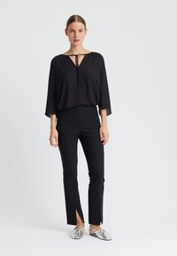 Schwarze Bluse mit V-Ausschnitt und Bindedetail, kombiniert mit schwarzen, schmal zulaufenden Hosen mit Seitenschlitzen. Silberne Slippers runden den Look ab.