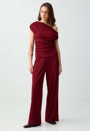 Kobieta stoi w teksturowanym ciemnoczerwonym topie z jednym ramieniem i dopasowanych szerokich spodniach, nosi złotą bransoletkę na ramieniu i ciemne buty.