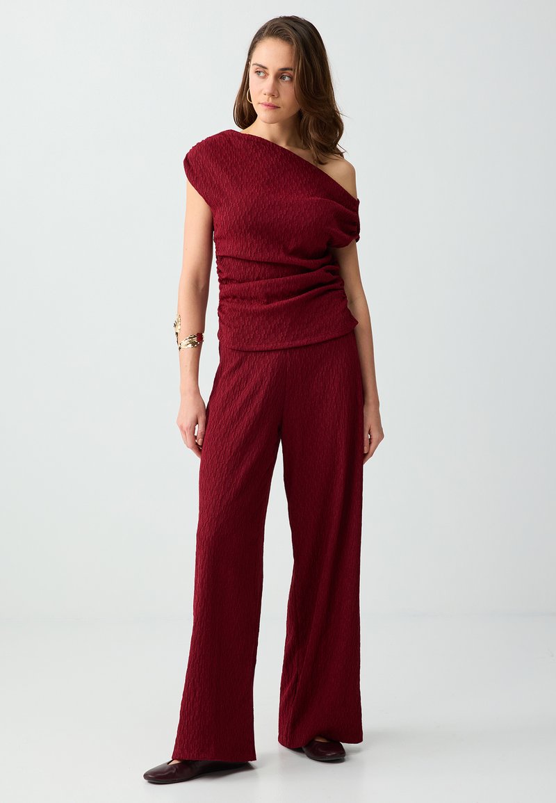 Femme debout avec un top rouge foncé texturé à une épaule et un pantalon large assorti, portant un bracelet doré au bras et des chaussures foncées.