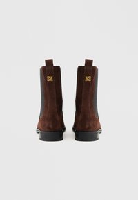 Barna szarvasbőr bokacsizma elasztikus oldalbetétekkel és arany díszítéssel, melyeken az "SM" és "M2" monogramok láthatók. A design-t sík fekete talpak teszik teljessé.