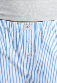 Boxer shorts de algodón a rayas verticales en azul y blanco, con cinturilla elástica y dos botones blancos en la parte frontal, llevados bajo una camiseta de canalé gris.