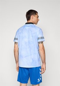 Maillot bleu clair à manches courtes avec un col noir et des accents à motifs sur les manches. Porté avec un short bleu mettant en avant un logo et un design blancs.
