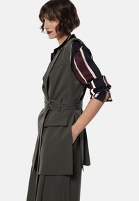 Donna con capelli corti castani indossa un gilet grigio scuro con cintura sopra una maglietta a righe a maniche lunghe arrotolate, in posa di profilo.