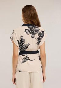 Blouse beige avec un motif floral noir, manches courtes, et une ceinture nouée à la taille marine. Tissu lisse avec une coupe décontractée et un ourlet arrondi.