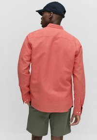 Koraalrood shirt met lange mouwen, knoopmanchetten en een rechte zoom, gecombineerd met olijfgroene shorts. De stof lijkt lichtgewicht en gestructureerd.