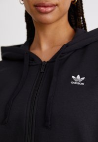 Juodas atsegamas gobtuvėlis, pagamintas iš minkštos medžiagos, su kengūros kišene ir siuvinėtu baltu "Adidas" logotipu kairiajame krūtinės pusėje.