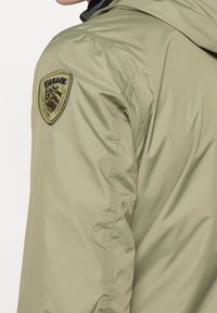 Chaqueta ligera verde con una textura suave, que presenta un parche cosido en el hombro izquierdo con un logo. Diseñada con capucha y cremalleras.