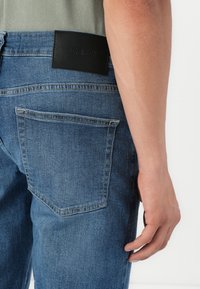 Acercamiento de la mano de una persona descansando junto a un par de jeans de mezclilla azules con una etiqueta negra en la cintura que dice "ONLY & SONS".