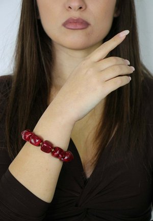 Czerwona masywna bransoletka wykonana z błyszczących koralików o różnych kształtach, z dodatkami w kolorze złotym. Noszona na jasnej skórze ręki.
