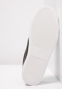 Baskets noires avec une tige texturée et une semelle en caoutchouc blanche. Présentent un logo embossé sur la semelle. Le dessous affiche un motif côtelé horizontal.