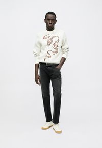 Beli pleteni pulover z rjavo-rjavo abstraktno obliko, kombiniran s črnimi jeans hlačami in belimi supergami z gumijastim podplatom.