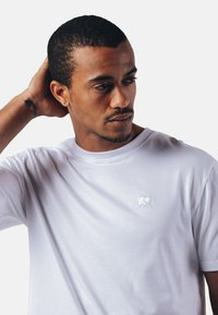 T-shirt en coton blanc avec un logo d'éléphant brodé discret sur la poitrine, col rond et manches courtes. Texture lisse.