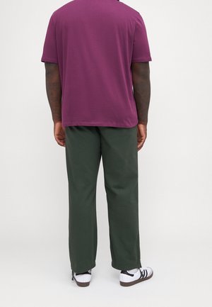 Mężczyzna w ciemnozielonych spodniach, białych trampkach z czarnymi paskami i fioletowej koszulce z krótkim rękawem stoi tyłem na jednolitym tle.