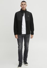 Svart fleecejakke med en glatt overdel, høy krage og glidelås foran. Kombinert med en hvit t-skjorte og grå slim-fit jeans. Svart sneakers.