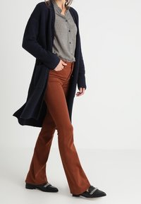 Dlouhý námořnický kardigan, kostkovaná košile na knoflíky, rezavě zbarvené rozšířené kalhoty a černé mokasíny s kovovými doplňky. Materiál má texturovaný pletený vzhled.