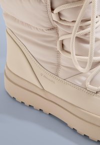 Beige geïsoleerde laars met een gevoerde bovenkant, gestructureerde veters en een bolle zool met een duidelijk profiel. "Primaloft"-merk aanwezig.