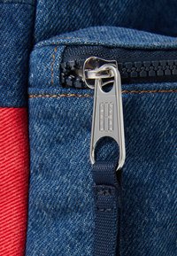 Sac à dos en denim avec une touche rouge, des accessoires en métal argenté, et une sangle navy. Tissu texturé avec une finition légèrement usée.