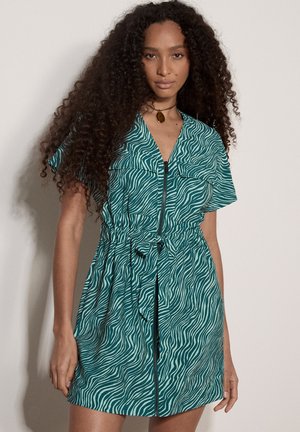 Frau mit langen lockigen Haaren, die ein kurzärmliges türkis-weißes Zebra-Muster-Kleid mit Frontreißverschluss und Taillengürtel trägt, steht vor einer schlichten Wand.