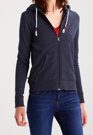 Mujer con sudadera con capucha gris oscuro con cierre y cordones blancos, jeans azules y camiseta roja visible debajo de la sudadera.