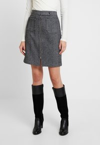 Mini-jupe noir et blanc en écaille de poisson avec fermeture éclair à l'avant, deux poches et coupe droite, associée à des bottes noires à talons hauts jusqu'aux genoux.