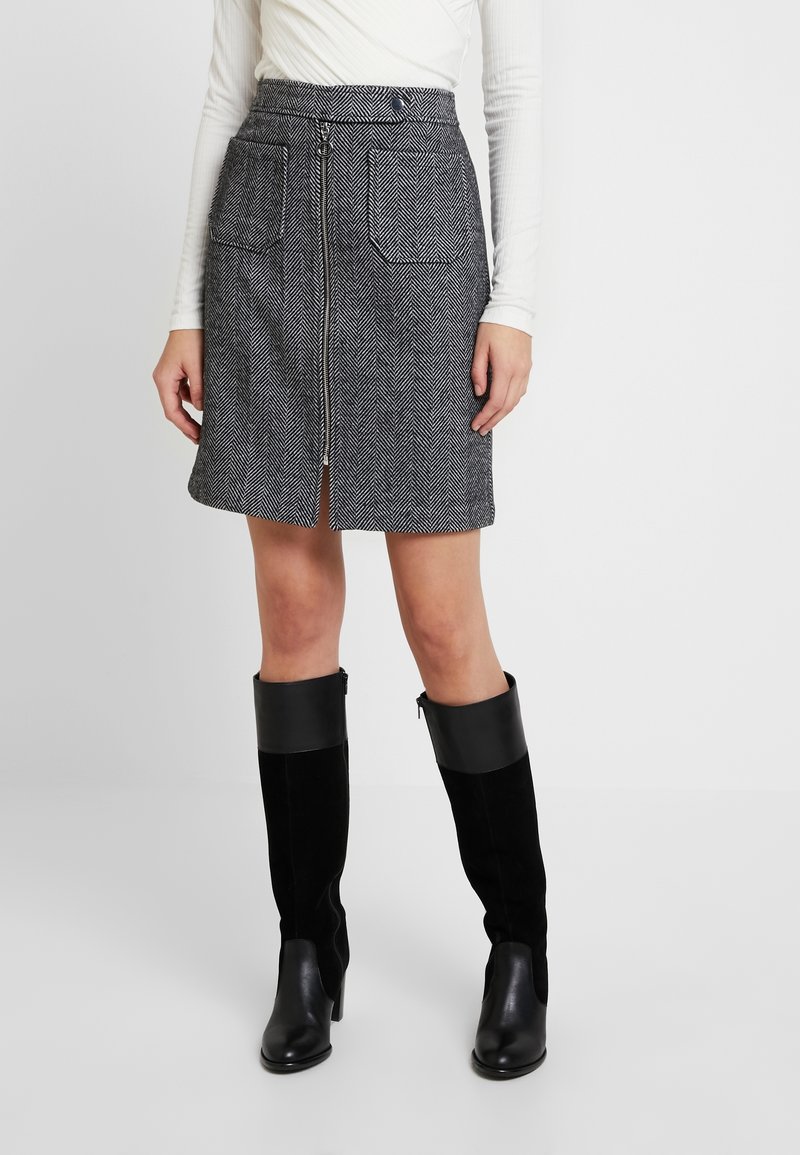 Mini-jupe noir et blanc en écaille de poisson avec fermeture éclair à l'avant, deux poches et coupe droite, associée à des bottes noires à talons hauts jusqu'aux genoux.