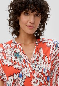 Lebhaftes oranges Hemd mit floralem Muster, mit Kragen und Knopfdetails, aus leichtem Stoff mit glatter Textur.