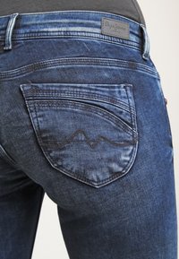 Mörkblå denimjeans med en distinkt fickdesign som har vågigt sömnad. Metallhardware och texturerade tygdetaljer är synliga.