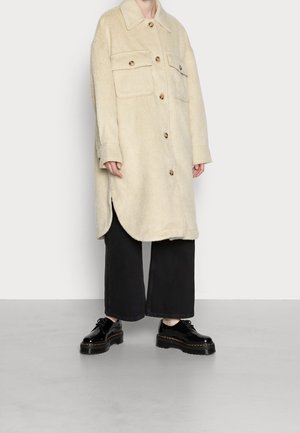 Person iført en lang beige knappet frakke med klaplommer, brede sorte korte bukser og sorte blanke snørestøvler med plateau.