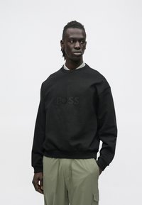 Άνδρας μοντέλο που φοράει μαύρο φούτερ με διακριτικό λογότυπο "BOSS" και παντελόνι χρώματος ελιάς, στέκεται μπροστά σε απλό φωτεινό φόντο.