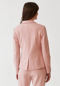Světle růžový blazer s upraveným střihem, s klopovým límcem a jedním rozparkem vzadu. Hladká tkanina s jemnou texturou.