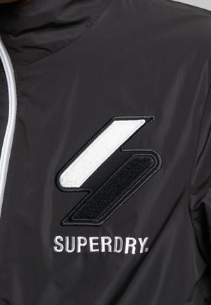 Chaqueta negra con un destacado logotipo bordado en blanco y negro con un acabado texturizado. Diseño con cremallera frontal y cuello alto.