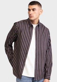 Cămășă cu dungi, în combinația de culori albastru marin și ruginiu, cu linii verticale, guler standard și croială lejeră, purtată peste un tricou alb.