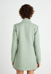 Abrigo verde claro con un tejido texturizado y suave. Presenta mangas largas y un cuello alto, acabando por encima de la rodilla, con una silueta ajustada.