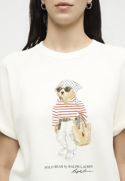 Αρκούδα με γυαλιά ηλίου, ριγέ πουκάμισο, λευκό παντελόνι, μαντήλι στο κεφάλι, κρατάει μπεζ τσάντα tote με το λογότυπο Polo σε λευκό φούτερ.