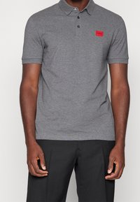 Camisa polo gris con un cuello clásico, mangas cortas y un parche de logo rojo en el lado izquierdo del pecho; tejido suave con una textura lisa.
