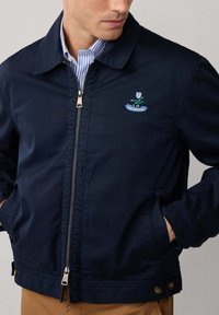 Uomo che indossa una giacca a zip blu navy con un emblemma ricamato sul petto, una camicia a righe blu sotto, e pantaloni marroni, con le mani in tasca.