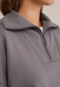 Grå zip-up sweatshirt med høj krave, der har en metal lynlås og struktureret stof. Nærbillede med fokus på kraven og lynlåsdetaljen.