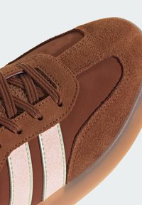 Brązowe zamszowe sneakersy z teksturowanym materiałem, jasnoróżowymi akcentami i trzema białymi paskami. Zaprojektowane z gładką gumową podeszwą dla lepszej przyczepności.