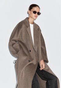 Dubbelzijdige jas van lichtbruin wolmengsel, met oversized schouders en zwarte knopen. Gekenmerkt door een witte shirt en donkere spijkerbroek.